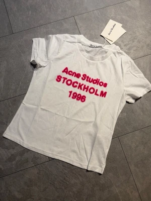 Vit Acne Studios t-shirt med tryck M - Säljer en vit t-shirt från Acne Studios med rosa text 'Acne Studios STOCKHOLM 1996' på bröstet. Klassisk rund halsringning och korta ärmar. Perfekt statement-plagg för dig som gillar stilrena men ändå iögonfallande detaljer.