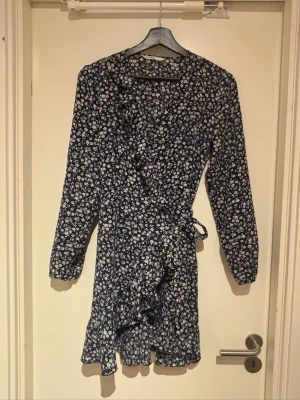 Blommig omlottklänning med volang - Mörkblå kort omlottklänning med vitt blommönster och volangdetaljer längs kanten. Klänningen har långa ärmar och knyt i midjan för en snygg passform. Perfekt för vår och sommar! (XS-S)