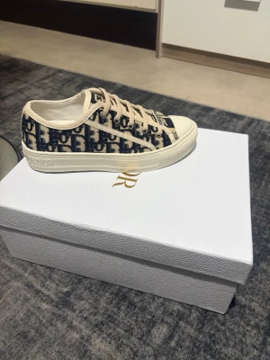 Dior Oblique låga sneakers - Snygga låga sneakers från Dior med klassiskt blått och beige Oblique-mönster. Skorna har vita sulor med Dior-logga och svart stjärndetalj undertill. Ovandelen är i canvas med beige detaljer och matchande snörning. Perfekt för dig som gillar lyxig streetstyle.