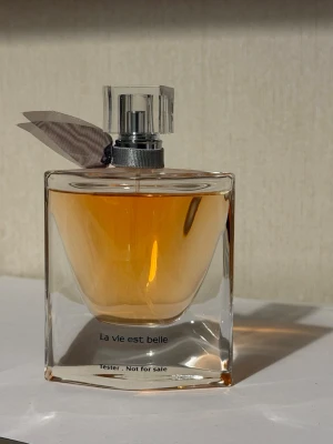 La vie est belle parfymflaska - Elegant parfymflaska med texten 'La vie est belle'. Flaskan är genomskinlig med en varm bärnstensfärgad vätska och har ett dekorativt band runt halsen. Locket är i klar plast och silverfärgad metall. Modern och stilren design som passar perfekt på sminkbordet.