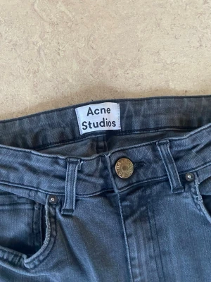 Grå jeans Acne Studios - Snygga grå jeans från Acne Studios med klassisk femficksmodell och raka ben. Jeansen har hög midja och är tillverkade i ett mjukt denimtyg som ger en avslappnad men stilren look. Det finns två brister, det är ett litet hål vid bälteshållaren, (lagat) så det syns inte så mycket. Det är slitet ovanför högra bakfickan men syns inte heller om man inte kollar noggrant. Storlek 29/30
