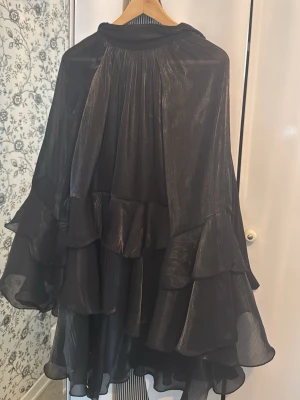 Svart volangklänning med v-ringning - Säljer en svart kortklänning med flera lager volanger och v-ringning. Klänningen har långa, vida ärmar och är gjord i ett glansigt, tunt material som ger ett elegant intryck. Perfekt för dig som gillar statement-plagg med mycket rörelse.