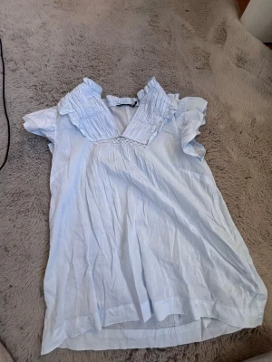 Ljusblå blus med volangkrage XS - Söt ljusblå blus från 157 i storlek XS. Blusen har korta ärmar, volangdetaljer vid kragen och ett lätt, luftigt tyg. Perfekt för en avslappnad och trendig look. Passar dig som gillar romantiska detaljer och en feminin stil.