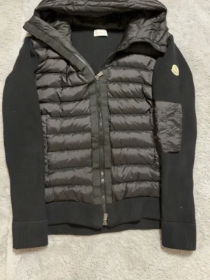 Svart cardiganjacka från Moncler - Snygg svart cardiganjacka från Moncler med quiltad framsida och huva. Jackan har ribbade muddar, dragkedja framtill och Moncler-logga på ärmen. Perfekt för lager-på-lager och stilrena outfits.