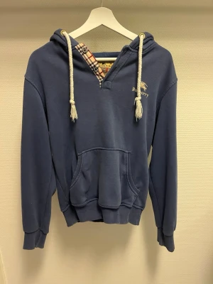 Blå burberry hoodie - Blå hoodie från Burberry med vita snören i huvan och klassiskt rutigt foder i halsen. Hoodien har känguruficka framtill och ribbade muddar vid ärmar och nederkant. Perfekt för en chill och stilren look. Den är storlek S men har krympt lite och sitter nu mer som xs