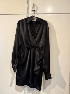 Svart omlottklänning från Zara - Snygg svart omlottklänning från Zara i glansigt material. Klänningen har lång ärm, krage och en elegant omlottdesign som ger en cool och stilren look. Perfekt för dig som gillar minimalistisk stil och vill ha något enkelt men ändå trendigt.