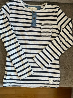 Randig långärmad topp från L.O.G.G./H&M - Snygg långärmad topp från L.O.G.G. med marinblå och vita ränder. Toppen har en liten bröstficka, två knappar framtill och detaljer i denim vid knäppningen. Perfekt till jeans eller kjol för en avslappnad look.