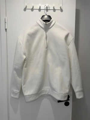 Vit halfzip hoodie - Säljer en stilren vit hoodie med halfzip och hög krage. Hoodien har långa ärmar, ribbade muddar och är tillverkad i ett mjukt sweatshirtmaterial. Perfekt för dig som gillar en clean och enkel look.