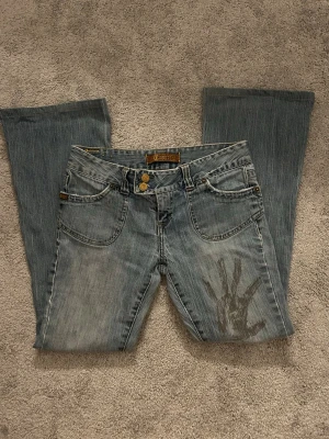 Ljusblå bootcut jeans med handavtryck - Säljer ett par ljusblå bootcut jeans med coolt handavtryck på ena benet och slitningar vid knät. Jeansen har klassisk femficksdesign, dubbla knappar i midjan och en avslappnad vintagekänsla. Perfekta för dig som gillar unik streetstyle.
