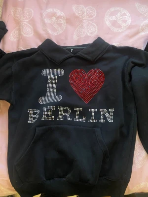 Svart hoodie med I ❤️ Berlin strass - Cool svart hoodie med texten 'I ❤️ BERLIN' i glittrande strass på bröstet. Hjärtat är gjort av röda stenar och resten av texten i silverfärgade stenar. Hoodien har magficka och ribbade muddar. Perfekt för dig som gillar statement-plagg och vill sticka ut.
