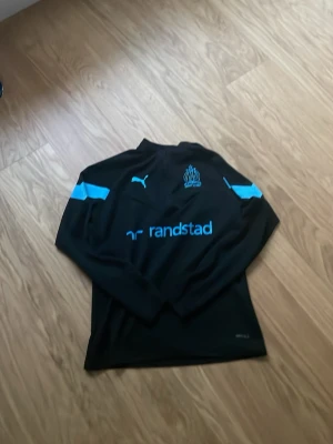 Svart långärmad Marseille träningströja - Svart långärmad träningströja från Puma med ljusblå detaljer och Olympique de Marseille-logga på bröstet. Tröjan har tryckt sponsorlogga och är tillverkad i ett funktionsmaterial som passar perfekt för sport och träning.
