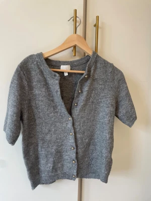 Grå kortärmad kofta från H&M - Mysig grå kofta från H&M med korta ärmar och rund hals. Koftan har knappar framtill i metall och är stickad i mjukt material. Perfekt att slänga över en topp för en chill look.