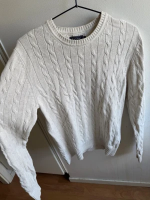 Beige kabelstickad tröja - Kabelstickad tröja beige med rund halsringning och ribbade muddar vid ärmslut och nederkant. Klassisk och stilren design som passar till det mesta. Tröjan har ett snyggt kabelmönster över hela plagget och är gjord i ett mjukt material.