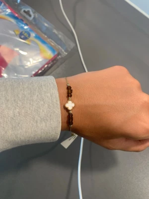 Guldigt armband med bruna pärlor - Snyggt armband med tunnt guldigt band, dekorerat med bruna pärlor och en fyrklöverformad detalj i vitt och guld. Helt nytt🌸