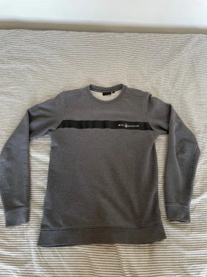 Grå sweatshirt med svart detalj - Snygg grå sweatshirt med svart band över bröstet och tryckt logga. Tröjan har rund halsringning, långa ärmar och ribbade muddar vid ärmslut och nederkant. Perfekt för en avslappnad och stilren look. Märket i nacken är lite sönder! 
