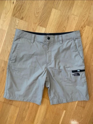 Beige shorts från The North Face (NYA!) - Helt nya beige shorts från The North Face med klassisk passform. Shortsen har bälteshällor, sidofickor och en praktisk ficka med dragkedja på benet där loggan är broderad. Perfekta för sommardagar och äventyr utomhus. Absolut nyskick! Kolla i storlekstabellen så storleken stämmer. 