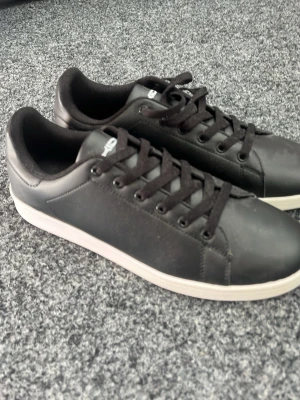 Svarta sneakers med vit sula - Stilrena svarta sneakers med vit sula och svarta skosnören. Ovandelen är i ett slätt material och designen är klassisk med lågt skaft. Passar perfekt till jeans eller shorts för en clean look.