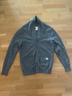 Grå kofta från Jack & Jones - Mysig grå kofta från Jack & Jones med sjalkrage och knäppning framtill. Två framfickor och ribbade muddar vid ärmslut och nederkant. Liten patch på ena fickan för extra detalj. Perfekt för lager på lager och chill stil.