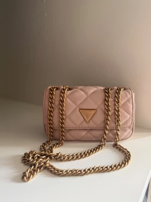 Rosa quiltad axelväska från Guess - Supersnygg rosa axelväska från Guess med quiltat mönster och guldfärgad kedjerem. Väskan har en klassisk rektangulär form och Guess-logga i guld på framsidan. Tillverkad i konstläder som känns mjukt och lyxigt. Perfekt för dig som vill ha en trendig och stilren väska.