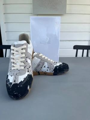 Maison Margiela svartvit sneakers - Unika sneakers från Maison Margiela med vit och grå ovandel i läder och mocka, samt svart färgstänksmönster över hela skon. Klassisk gummisula och vita snören ger en clean men edgy vibe. Perfekt för dig som vill sticka ut med din stil.