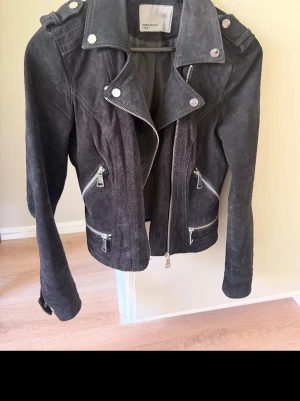 Svart mockajacka från Vero Moda - Snygg svart mockajacka från Vero Moda med silvriga dragkedjor och detaljer. Jackan har klassisk bikerstil med bred krage, axelklaffar och flera fickor med dragkedja. Perfekt för dig som vill ha en cool och tidlös look.