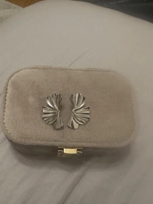 Silvriga örhängen i veckad design - Säljer ett par statement örhängen i silverfärgad metall med unik veckad form som påminner om ett vikt tyg eller blad. De är stora och täcker en del av örsnibben, vilket ger en cool och modern look. Perfekta för dig som vill sticka ut med dina accessoarer.