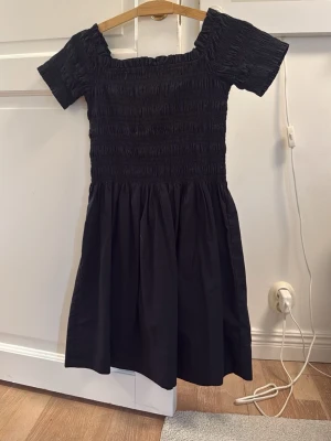 Svart smockad klänning från H&M - Supersöt svart klänning från H&M med smockad överdel och korta ärmar. Klänningen har en lätt utställd kjol och offshoulder-design som ger en trendig vibe. Perfekt för dig som gillar enkel men snygg stil.