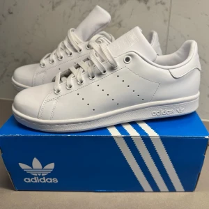 Vita Adidas Stan Smith sneakers - Klassiska helvita Adidas Stan Smith sneakers med lågt skaft och diskret perforerad design på sidorna. Skorna har vit sula, vita skosnören och är tillverkade i slätt läder. Perfekta för en clean och stilren look.