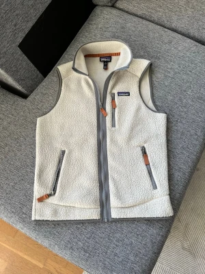 Vit fleeceväst från Patagonia - Snygg vit fleeceväst från Patagonia med grå detaljer och dragkedjor med bruna läderflärpar. Västen har hög krage, två sidofickor och en bröstficka. Perfekt för lager-på-lager och riktigt mysig med sitt fluffiga material.