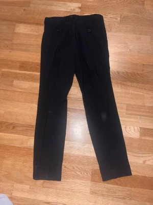 Svarta raka kostymbyxor - Snygga svarta kostymbyxor med rak passform och klassisk design. Byxorna har två bakfickor med knappar och är perfekta för en stilren look. Materialet ser ut att vara en blandning av polyester och viskos för en mjuk känsla.