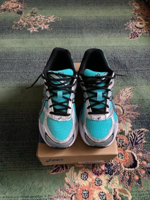 Turkosa och vita Asics sneakers - Snygga sneakers från Asics i turkos, vitt och svart med mesh-ovandel för bra ventilation. Klassisk Asics-logga på sidan och svarta skosnören. Sulan är vit med svarta detaljer och skon har en sportig, modern look som passar till många stilar.