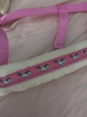 Rosa grimma med mjuk foder och hästar - Säljer en söt rosa grimma med mjuk vit fleecefodring och detaljer med tecknade hästar på bandet. Grimman har justerbara remmar och metallspännen. Perfekt för dig som vill ha något gulligt till din ponny eller häst. Storlek full shettis 