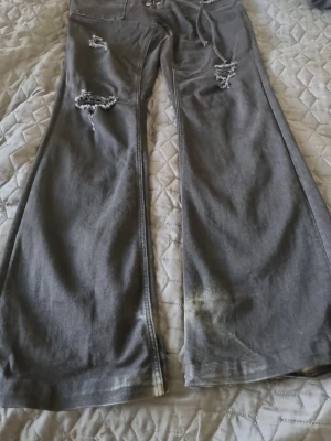 Svart vida jeans med slitningar - Säljer ett par gråa jeans med breda ben och slitna detaljer på låren och knäna. Jeansen har dragsko i midjan och en avslappnad passform. Perfekta för en chill och trendig look. Använd skick I storlek L 14+. Den är svart men ser ljusare ut på bilden på grund av kameran.  