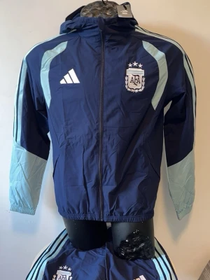 Blå Argentina adidas träningsjacka - Mörkblå träningsjacka med ljusblå detaljer från adidas och Argentina AFA-logga på bröstet. Jackan har huva, hel dragkedja och klassiska adidas-ränder längs ärmarna. Perfekt för fotbollsträning eller sportig stil.