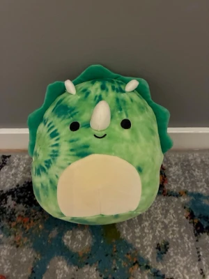 Grön dinosaurie Squishmallow - Supermjuk och rund grön Squishmallow i form av en dinosaurie med horn och vit mage. Den har ett batikmönster i ljus grön och mörk grönt samt söta detaljer som små öron och ett glatt ansikte. Perfekt som gosedjur eller dekoration i rummet.