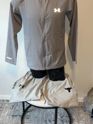 Grå träningsset från Under Armour - Säljer ett grått träningsset från Under Armour med jacka och byxor. Jackan har dragkedja och lång ärm, medan byxorna har elastisk midja och Project Rock-logga. Perfekt för gymmet eller löpning. Materialet är lätt och andas bra.
