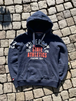 Mörkblå Superdry hoodie med tryck - Mörkblå hoodie från Superdry med vit och röd text på bröstet där det står 'Superdry State Athletics'. Stor huva med vita dragsnören och en klassisk känguruficka framtill. Mjuk bomullsblandning och ribbade muddar vid ärmar och nederkant.