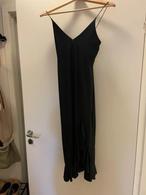 Svart långklänning från NLY One - Svart långklänning från NLY One med smala axelband och v-ringning. Klänningen har volangdetalj nertill och är tillverkad i ett glansigt, mjukt material som ger en elegant känsla. Perfekt för dig som vill ha en stilren och trendig look.