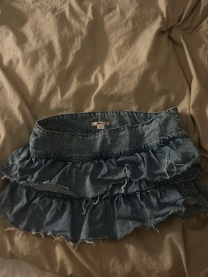 Blå volangkjol i denim från Gina Tricot andvänt en gång - Supersöt blå jeanskjol från Gina Tricot med volanger och råa kanter. Kjolen är kort och har en avslappnad vibe, perfekt till sneakers eller boots. Tillverkad i bomull för en skön känsla och snygg passform.
