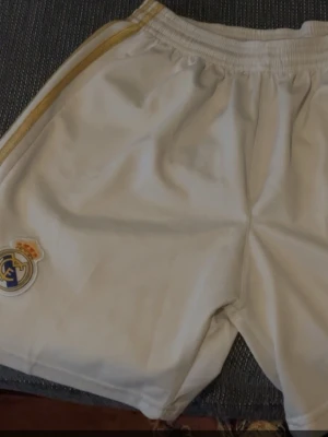 Vita Real Madrid shorts från Adidas - Säljer ett par vita Real Madrid shorts från Adidas med guldfärgade detaljer och klubbmärke på ena benet. Shortsen har elastisk midja och klassiska Adidas-ränder längs sidorna. Perfekta för fotboll eller träning.