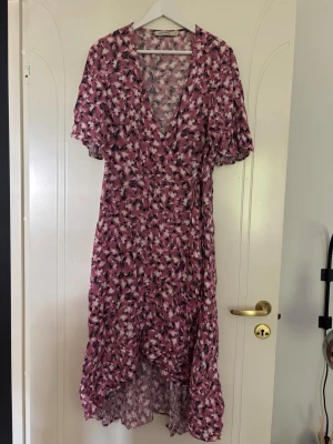 Rosa klänning från Carin Wester - Säljer en lång omlottklänning med blommigt mönster i rosa, vit och svart. Klänningen har v-ringning, volangkant nertill och korta ärmar. Perfekt för sommarens alla tillfällen och ger en romantisk look.