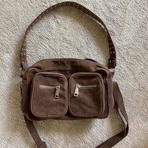 Mörkbrun axelväska - Brun axelväska från Noella i fint skick. Rymlig för att vara en axelväska med flertalet fack både på ytter- och innersidan. Kommer med två band varav det längre är avtagbart samt justerbart. Ca 25cm bred, 16cm hög samt 7 cm djup.