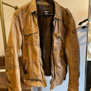 Tigha Läderjacka, M - En vintage brun läderjacka. Estimerad vara från runt 1980-talet. Bröstfickor med knapp och sidfickor med dragkedja. Finns 2 innerfickor som stängs med knapp. Den är i bra skick och charmigt sliten. Hämtas i Linköping, kan fraktas inom Sverige, frakt tillkommer.