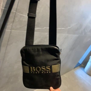 Hugo boss axel väska - En fin Hugo boss väska guldig och svart skick 6/10 lite slitet på guldiga texten