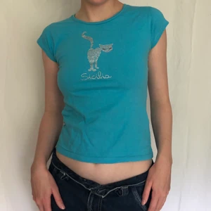 Turkos babytee - Fett gullig turkos babytee med en katt i rhinestones/strass från märket sicila, ögonen är rosa syns inte riktigt på bilden. Några stenar saknas annars bra skick. Stl xs-s. 