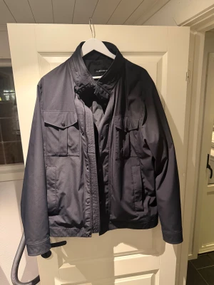 Svart vindjacka från J.Lindeberg - Stilren svart vindjacka från J.Lindeberg med klassisk krage, flera fickor med lock och dragkedja framtill. Jackan har en clean look och är perfekt för dig som klassisk stil. Storlek Xl men passar L med. Endast använd en gång. Ca 2 månader gammal. 