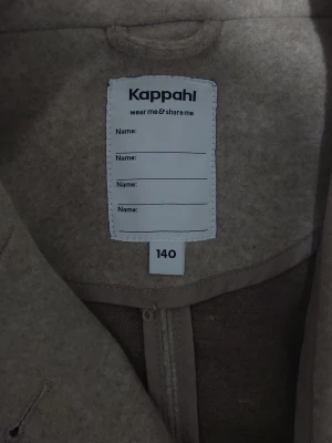 Beige kappa från Kappahl med knappar - Stilren beige kappa från Kappahl med klassisk rak passform och stora knappar framtill. Jackan har hög krage och är helfärgad, perfekt för dig som gillar en enkel och clean look. Passar till många olika outfits och har en tidlös design.