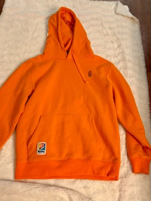 Orange hoodie från Coalition - Snygg orange hoodie från Coalition med stor ficka framtill och broderad logga på bröstet. Huva med dragsko och tryck på baksidan av huvan. Tillverkad i mjukt bomullsmaterial, perfekt för chill eller träning.