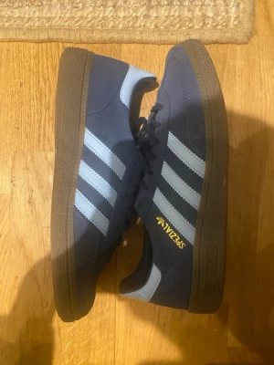 Adidas Spezial mörkblå sneakers - Säljer ett par Adidas Spezial sneakers i mörkblå mocka med ljusblåa klassiska tre ränder på sidorna och brun sula. Guldfärgad Spezial-logga på sidan och Adidas-mönstrad innersula. Skorna har snörning och retroinspirerad design.