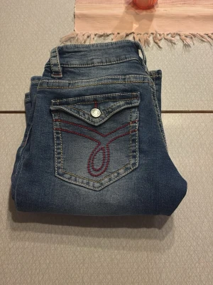Blå bootcut jeans med röd söm - Snygga blå jeans med bootcut-modell och cool röd dekorationssöm på bakfickan. Klassisk femficksdesign och knappstängning. Perfekta för dig som gillar en retroinspirerad look och vill sticka ut lite extra.